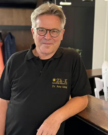 Dr. med. / med. dent. Arno König, medizinische Unterstützung und Longevity-Medizin, im schwarzen Poloshirt mit Zk-K-Logo, an eine Holztheke gelehnt