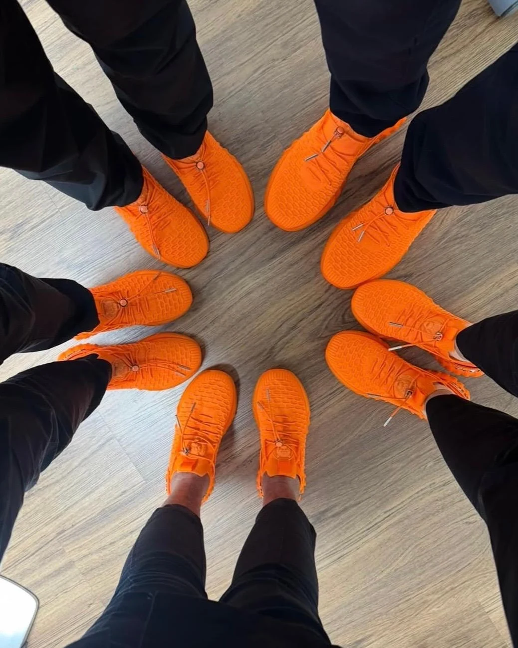 Füsse des Teams im Kreis, alle in gleichen orangen Turnschuhen und schwarzen Hosen, auf Parkett